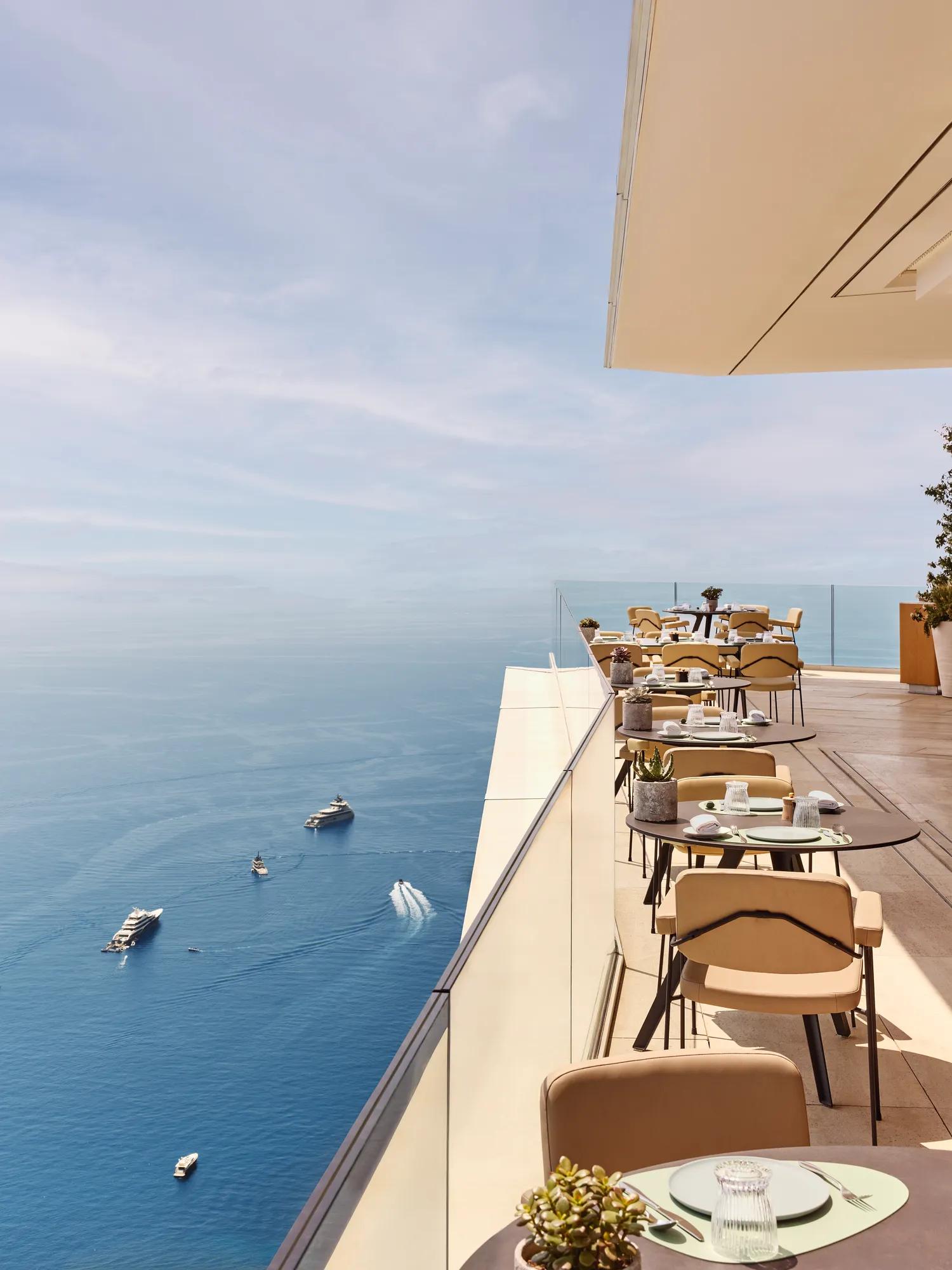 Restaurant en terrasse avec des tables élégamment dressées surplombant la Méditerranée, offrant une vue imprenable sur la mer et les yachts au loin.
