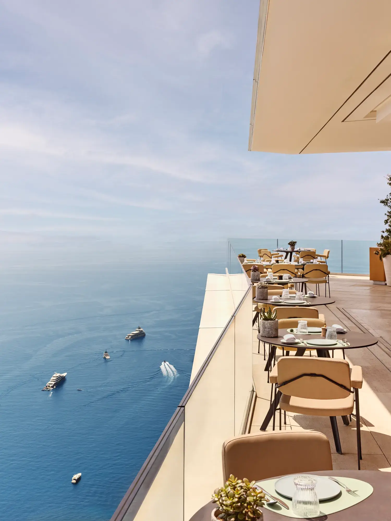 Terrace restaurant with elegantly set tables overlooking the Mediterranean, offering sweeping views of the sea with yachts in the distance.