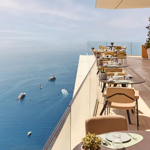 Restaurant en terrasse avec des tables élégamment dressées surplombant la Méditerranée, offrant une vue imprenable sur la mer et les yachts au loin.