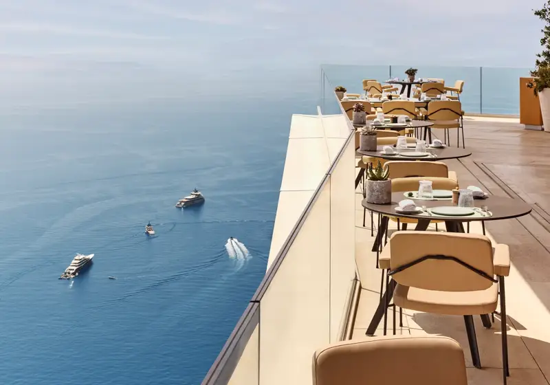 Restaurant en terrasse avec des tables élégamment dressées surplombant la Méditerranée, offrant une vue imprenable sur la mer et les yachts au loin.