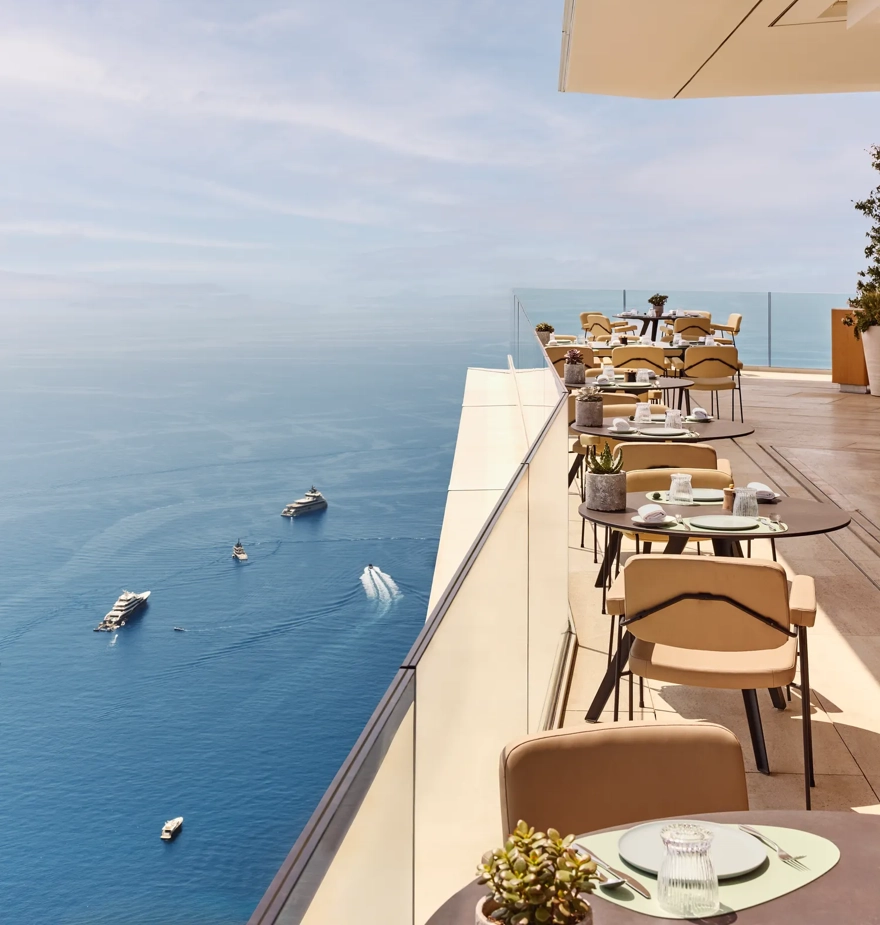Restaurant en terrasse avec des tables élégamment dressées surplombant la Méditerranée, offrant une vue imprenable sur la mer et les yachts au loin.