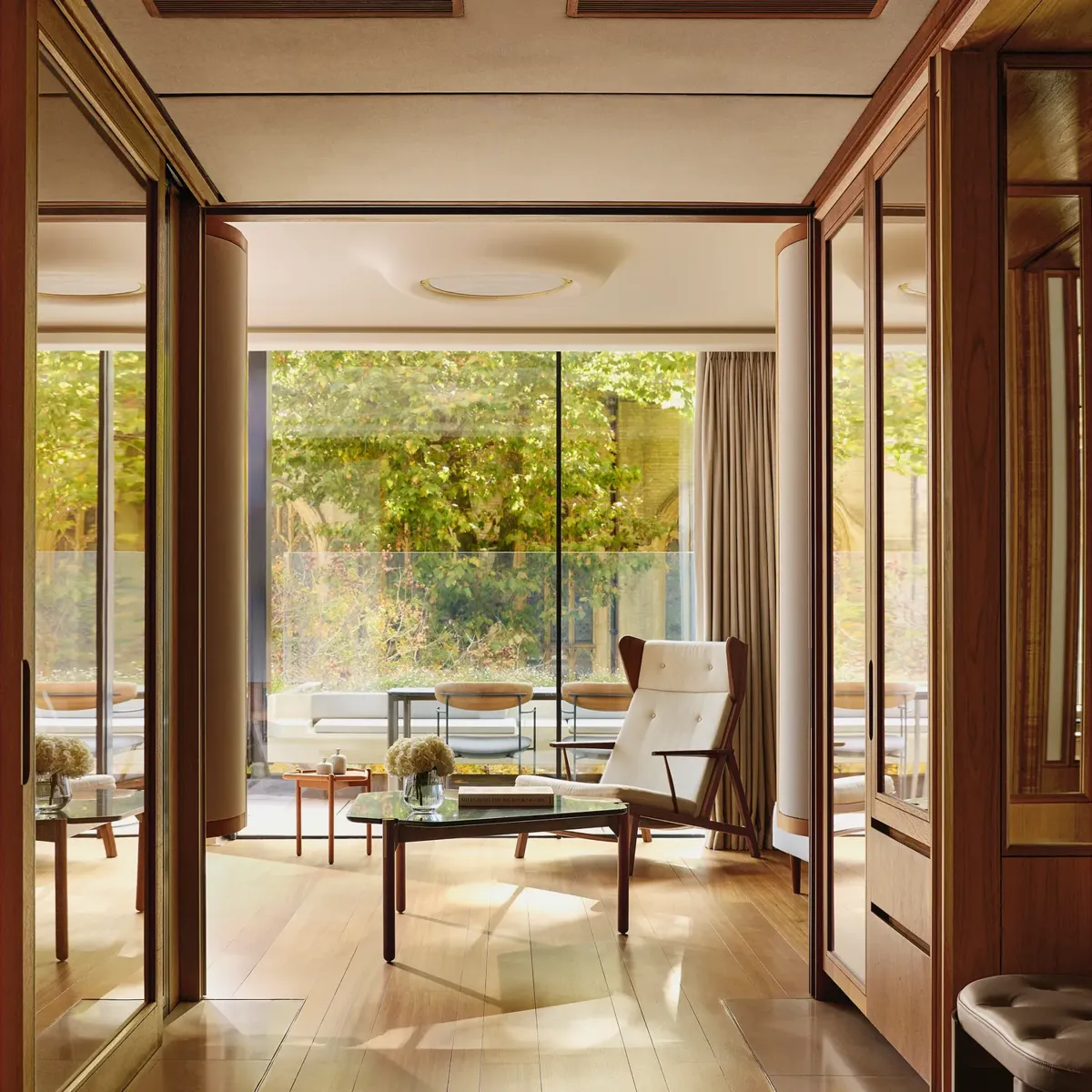 Light-filled suite with modern armchair, glass table, and terrace overlooking greenery, framed by floor-to-ceiling windows.