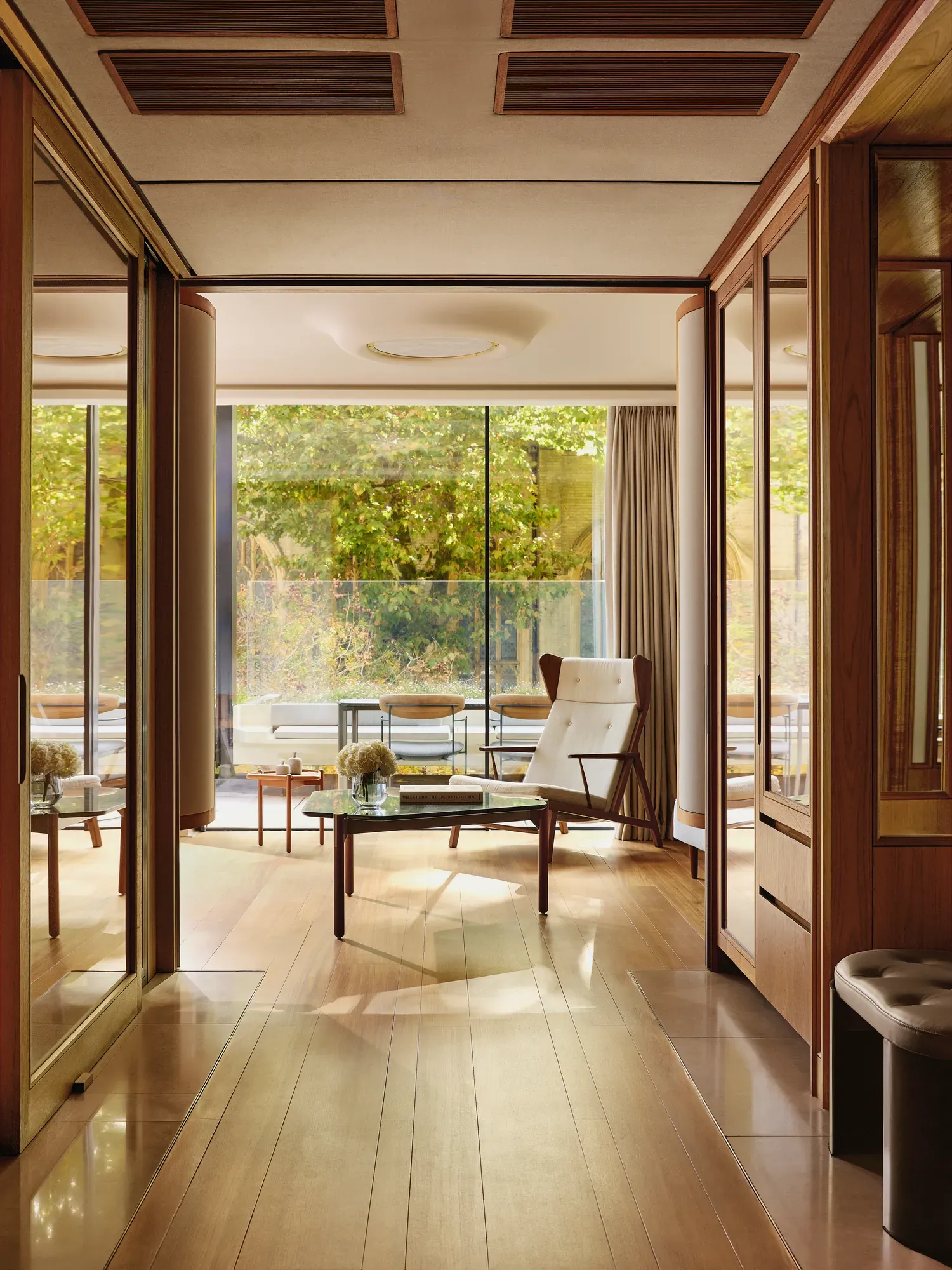 Light-filled suite with modern armchair, glass table, and terrace overlooking greenery, framed by floor-to-ceiling windows.