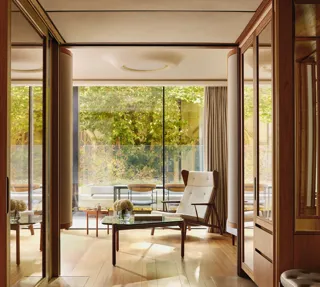 Light-filled suite with modern armchair, glass table, and terrace overlooking greenery, framed by floor-to-ceiling windows.
