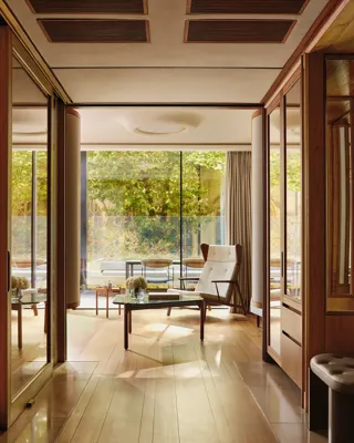 Light-filled suite with modern armchair, glass table, and terrace overlooking greenery, framed by floor-to-ceiling windows.