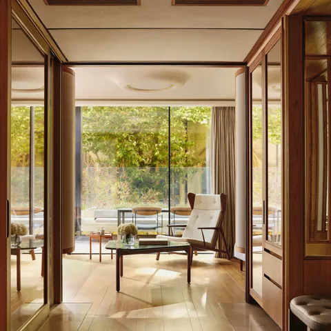 Light-filled suite with modern armchair, glass table, and terrace overlooking greenery, framed by floor-to-ceiling windows.