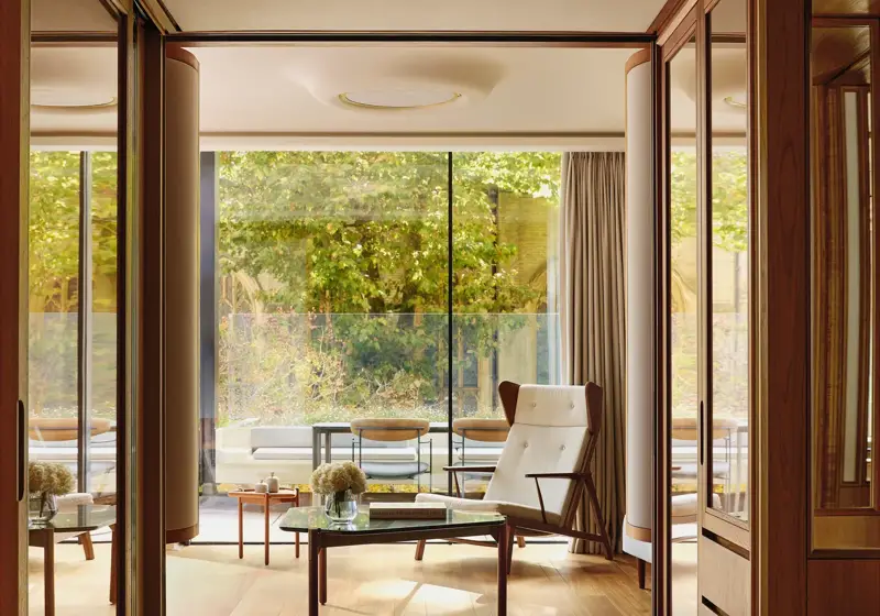 Light-filled suite with modern armchair, glass table, and terrace overlooking greenery, framed by floor-to-ceiling windows.