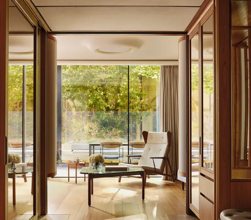 Light-filled suite with modern armchair, glass table, and terrace overlooking greenery, framed by floor-to-ceiling windows.