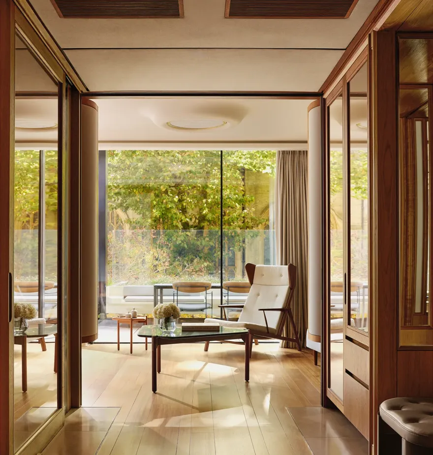 Light-filled suite with modern armchair, glass table, and terrace overlooking greenery, framed by floor-to-ceiling windows.