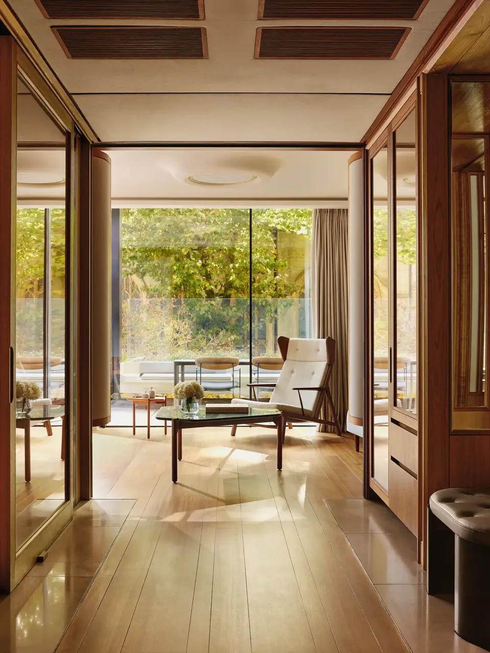 Light-filled suite with modern armchair, glass table, and terrace overlooking greenery, framed by floor-to-ceiling windows.