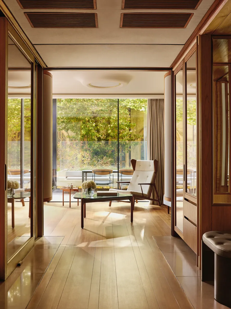 Light-filled suite with modern armchair, glass table, and terrace overlooking greenery, framed by floor-to-ceiling windows.