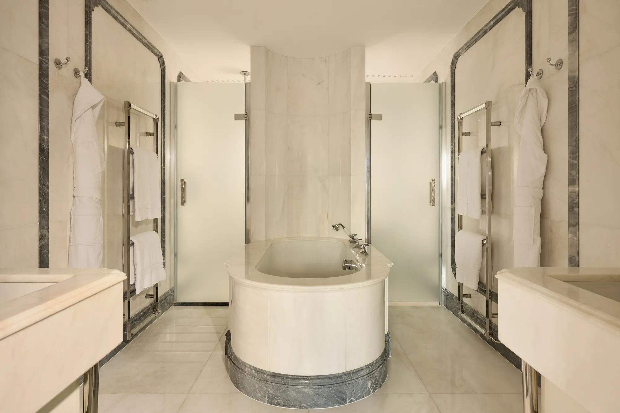 Luxurious marble bathroom with central oval bathtub, frosted glass doors, and towel rails.