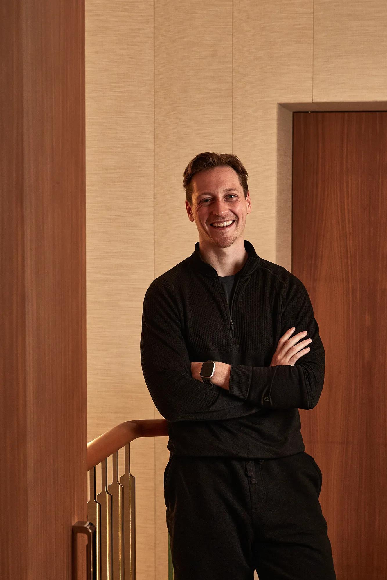 Smiling man in black sweater and trousers standing indoors with arms crossed.