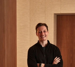 Smiling man in black sweater and trousers standing indoors with arms crossed.