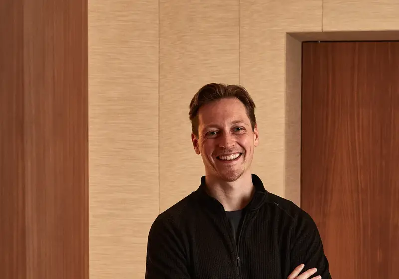 Smiling man in black sweater and trousers standing indoors with arms crossed.