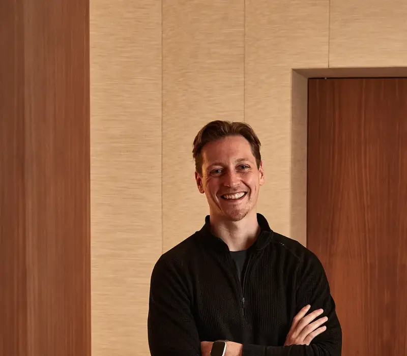 Smiling man in black sweater and trousers standing indoors with arms crossed.