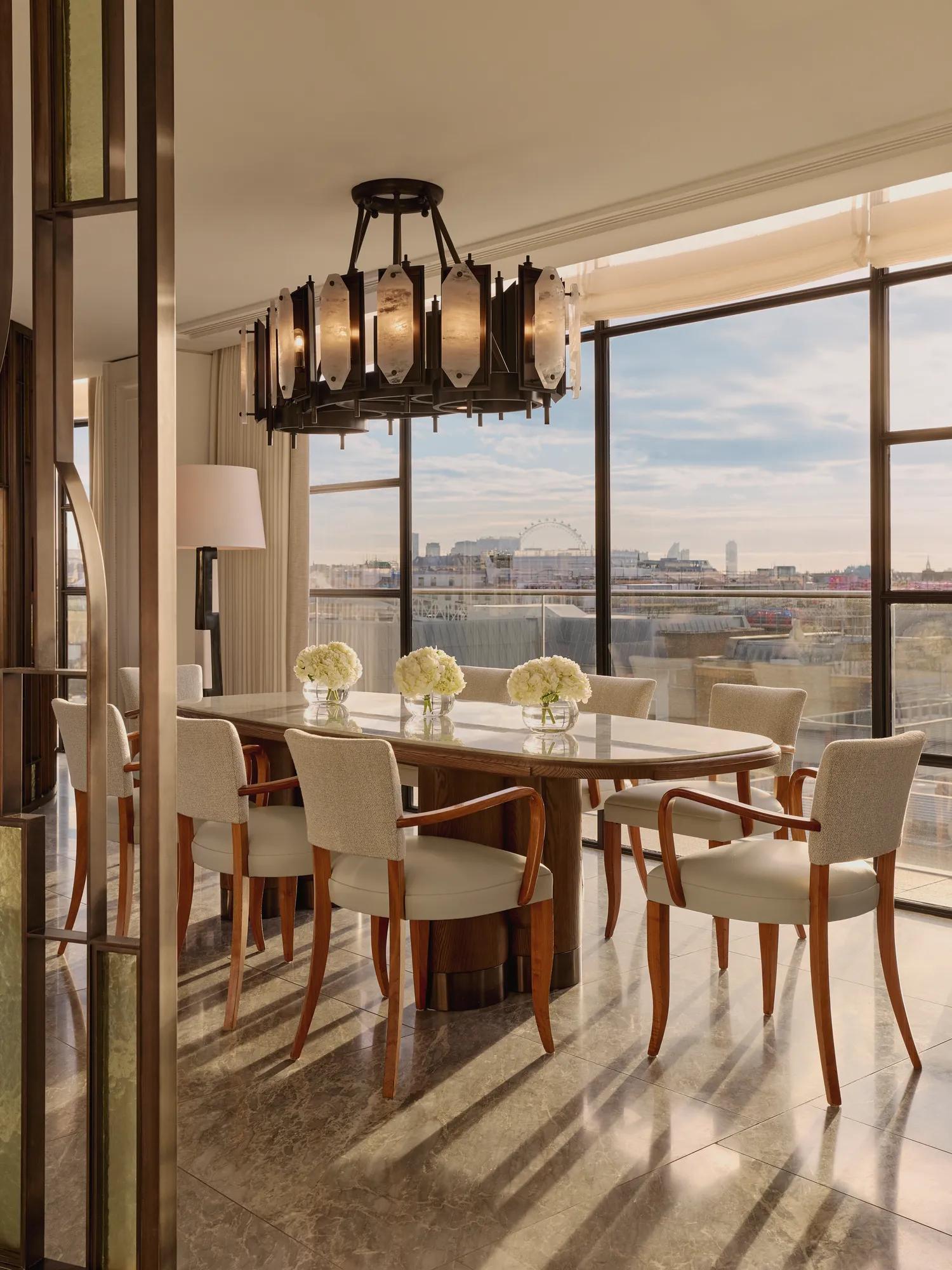 Elegant dining area with a long marble table surrounded by cream upholstered chairs, illuminated by a sculptural chandelier, with floor-to-ceiling windows revealing city views including the London Eye.
