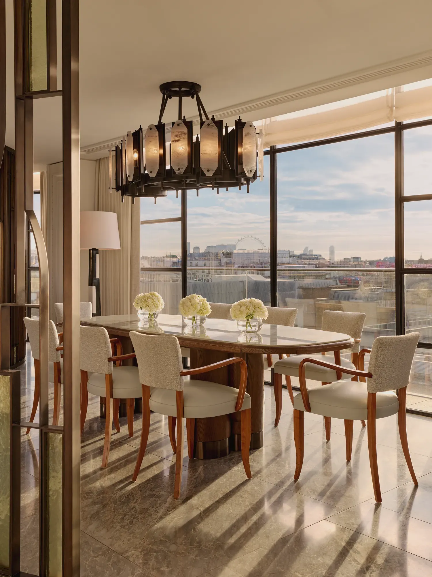 Elegant dining area with a long marble table surrounded by cream upholstered chairs, illuminated by a sculptural chandelier, with floor-to-ceiling windows revealing city views including the London Eye.