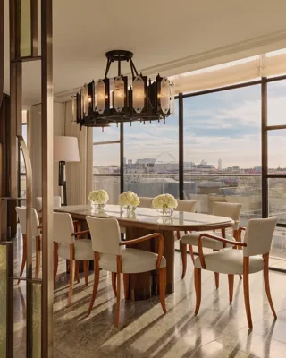 Elegant dining area with a long marble table surrounded by cream upholstered chairs, illuminated by a sculptural chandelier, with floor-to-ceiling windows revealing city views including the London Eye.