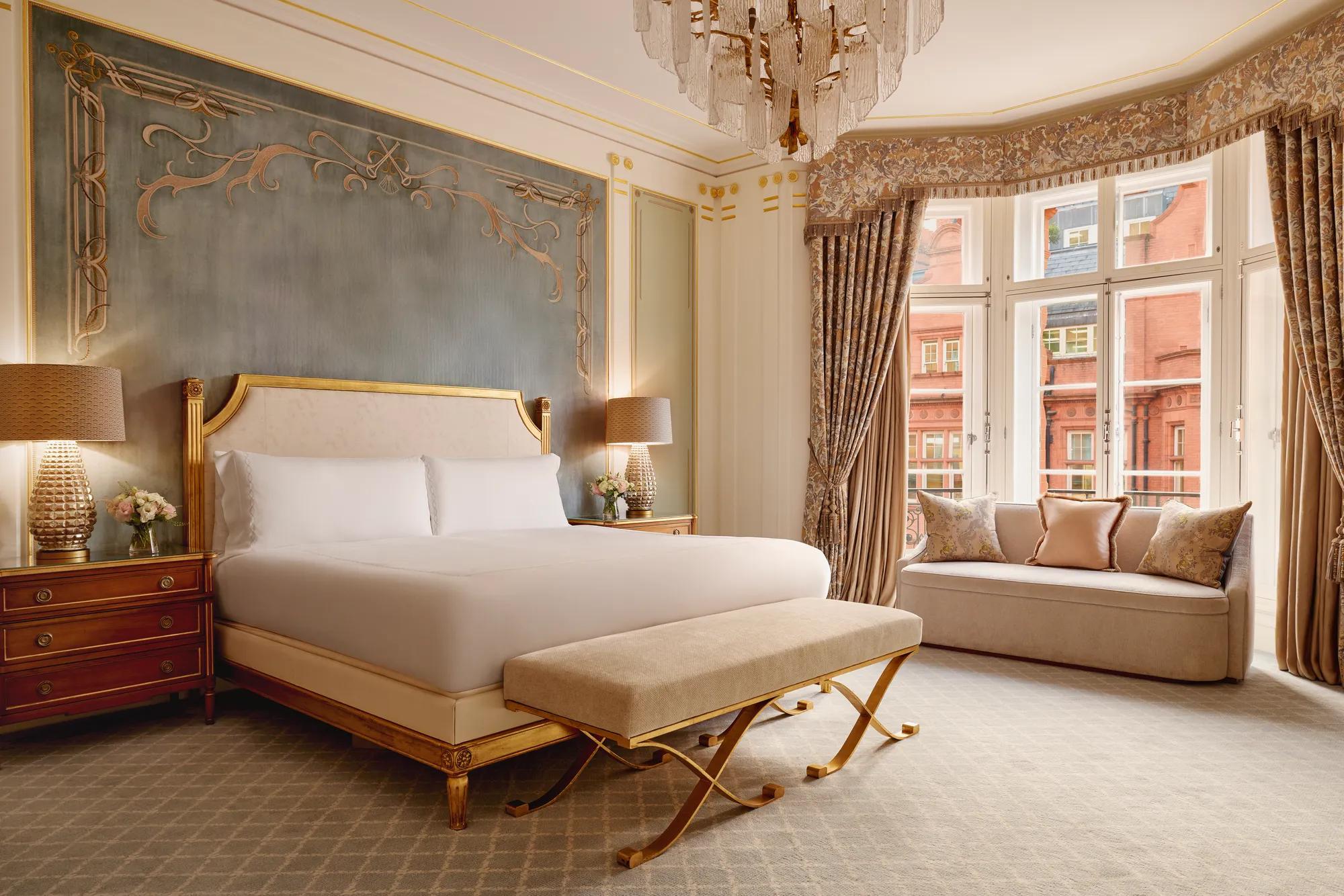 Elegant bedroom with a king bed framed by a gold headboard against a teal feature wall, two wooden bedside tables with lamps, and a window seat beneath tall curtained bay windows.