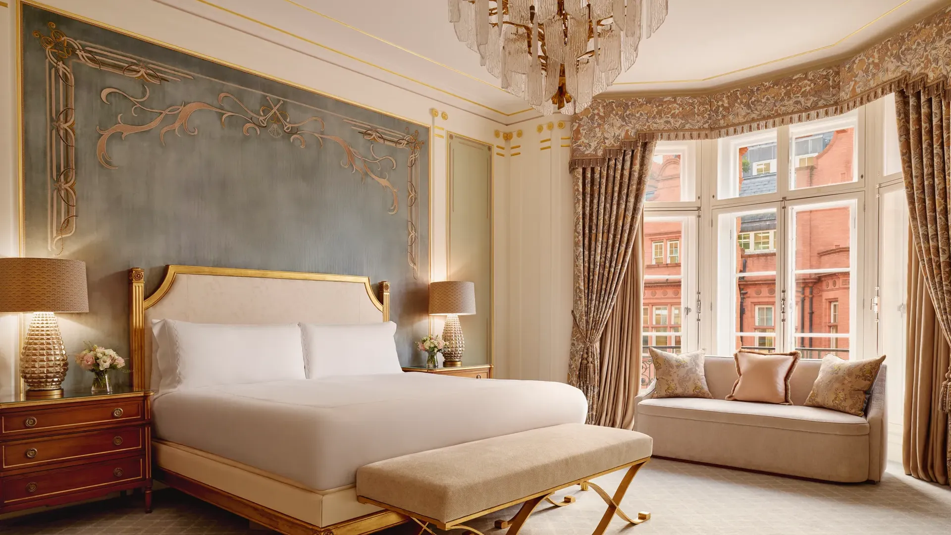 Elegant bedroom with a king bed framed by a gold headboard against a teal feature wall, two wooden bedside tables with lamps, and a window seat beneath tall curtained bay windows.