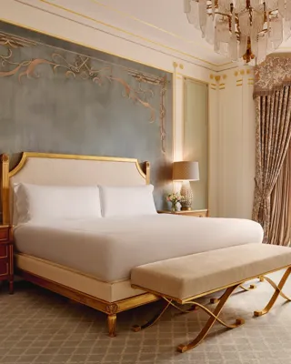 Elegant bedroom with a king bed framed by a gold headboard against a teal feature wall, two wooden bedside tables with lamps, and a window seat beneath tall curtained bay windows.