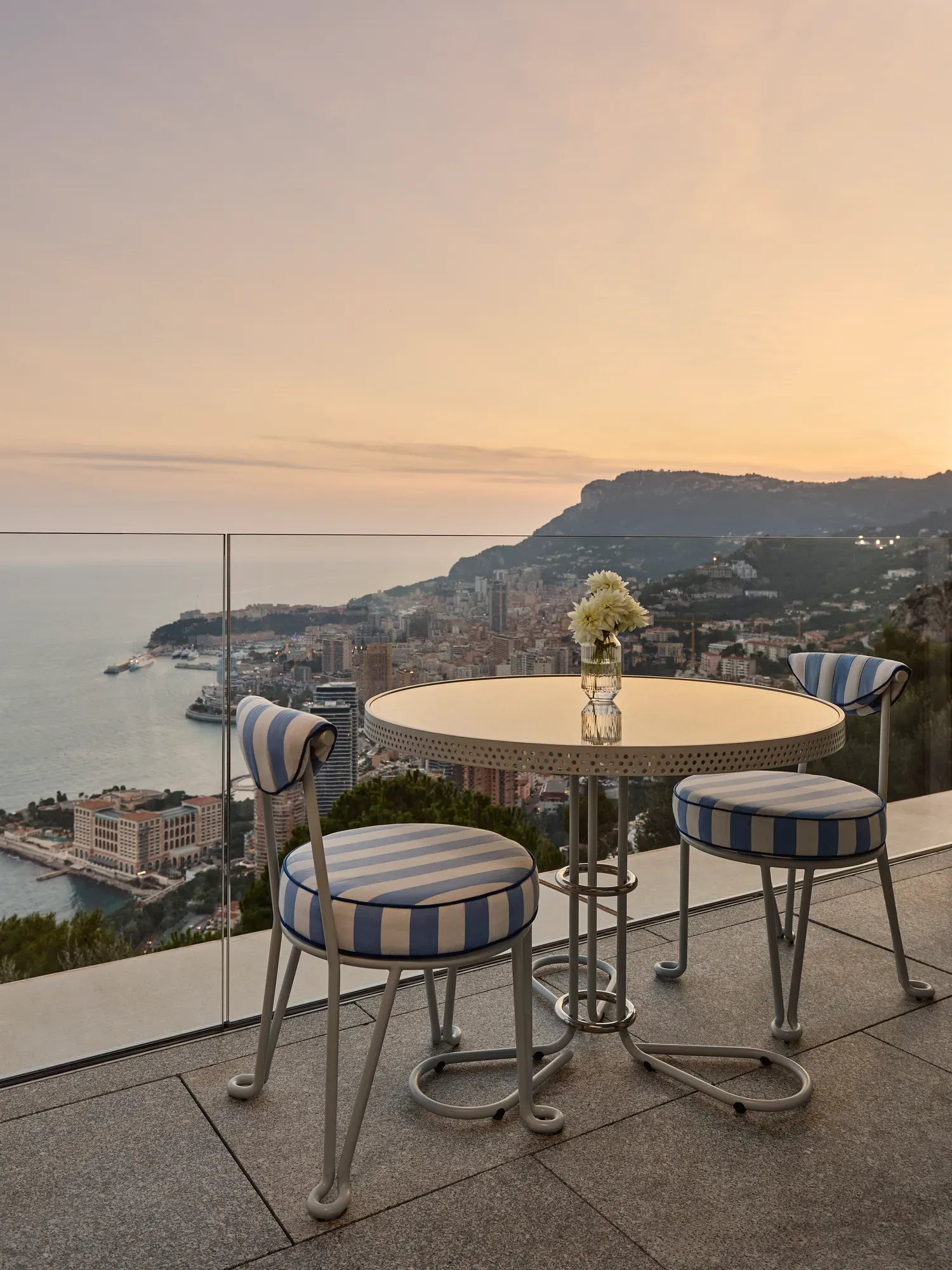 Une table ronde en métal avec un vase en verre contenant des fleurs blanches est posée sur une terrasse avec deux chaises aux coussins rayés bleu et blanc. La terrasse surplombe une ville côtière avec des bâtiments, des collines et la mer visibles en arrière-plan sous un ciel doux de coucher de soleil.