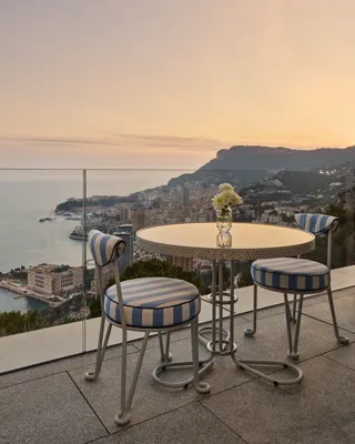 Une table ronde en métal avec un vase en verre contenant des fleurs blanches est posée sur une terrasse avec deux chaises aux coussins rayés bleu et blanc. La terrasse surplombe une ville côtière avec des bâtiments, des collines et la mer visibles en arrière-plan sous un ciel doux de coucher de soleil.