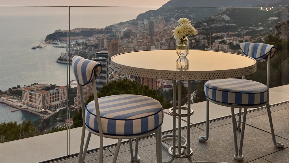 Une table ronde en métal avec un vase en verre contenant des fleurs blanches est posée sur une terrasse avec deux chaises aux coussins rayés bleu et blanc. La terrasse surplombe une ville côtière avec des bâtiments, des collines et la mer visibles en arrière-plan sous un ciel doux de coucher de soleil.