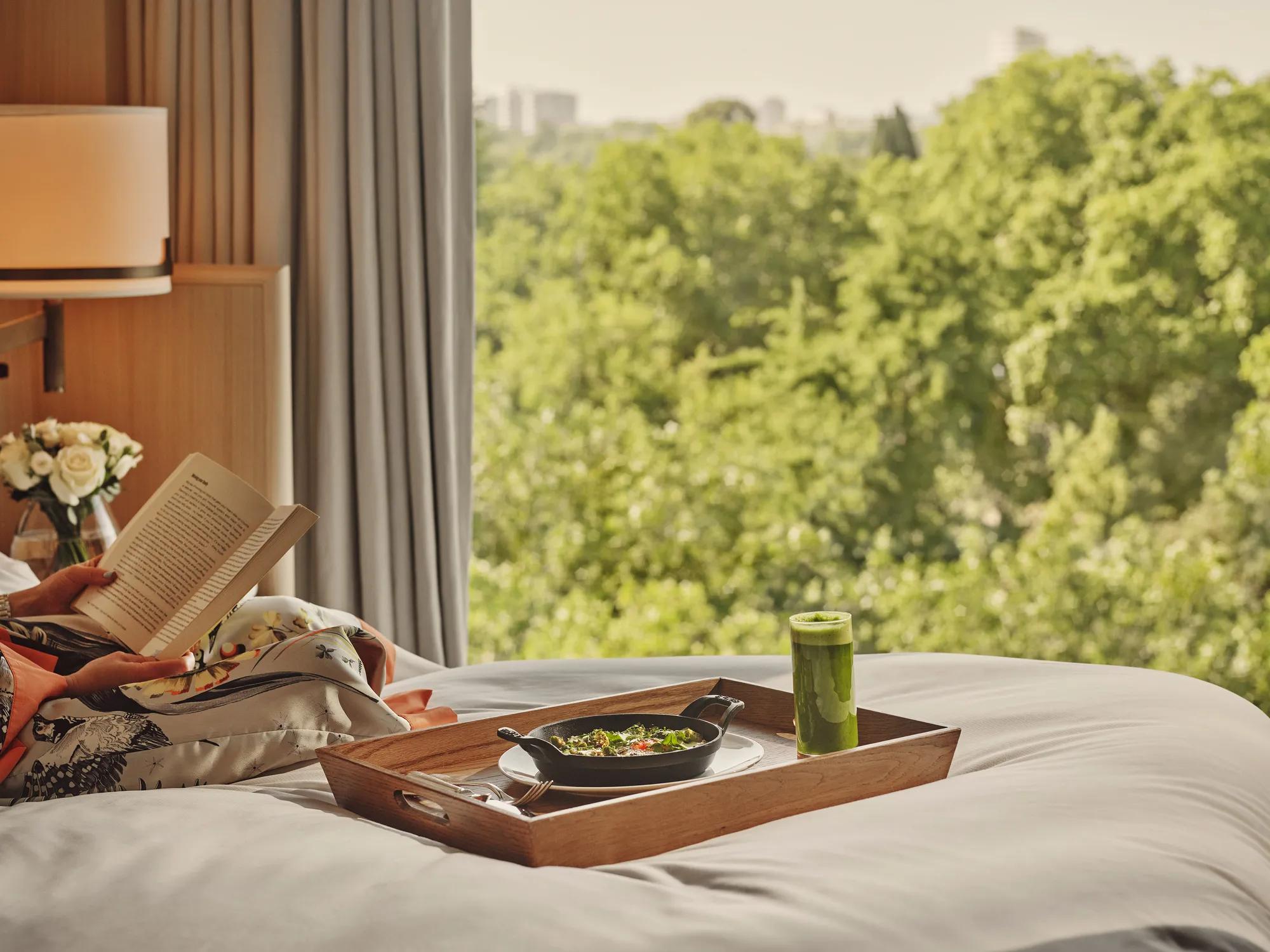 A breakfast tray with a green juice sitting on a bed overlooking greenery