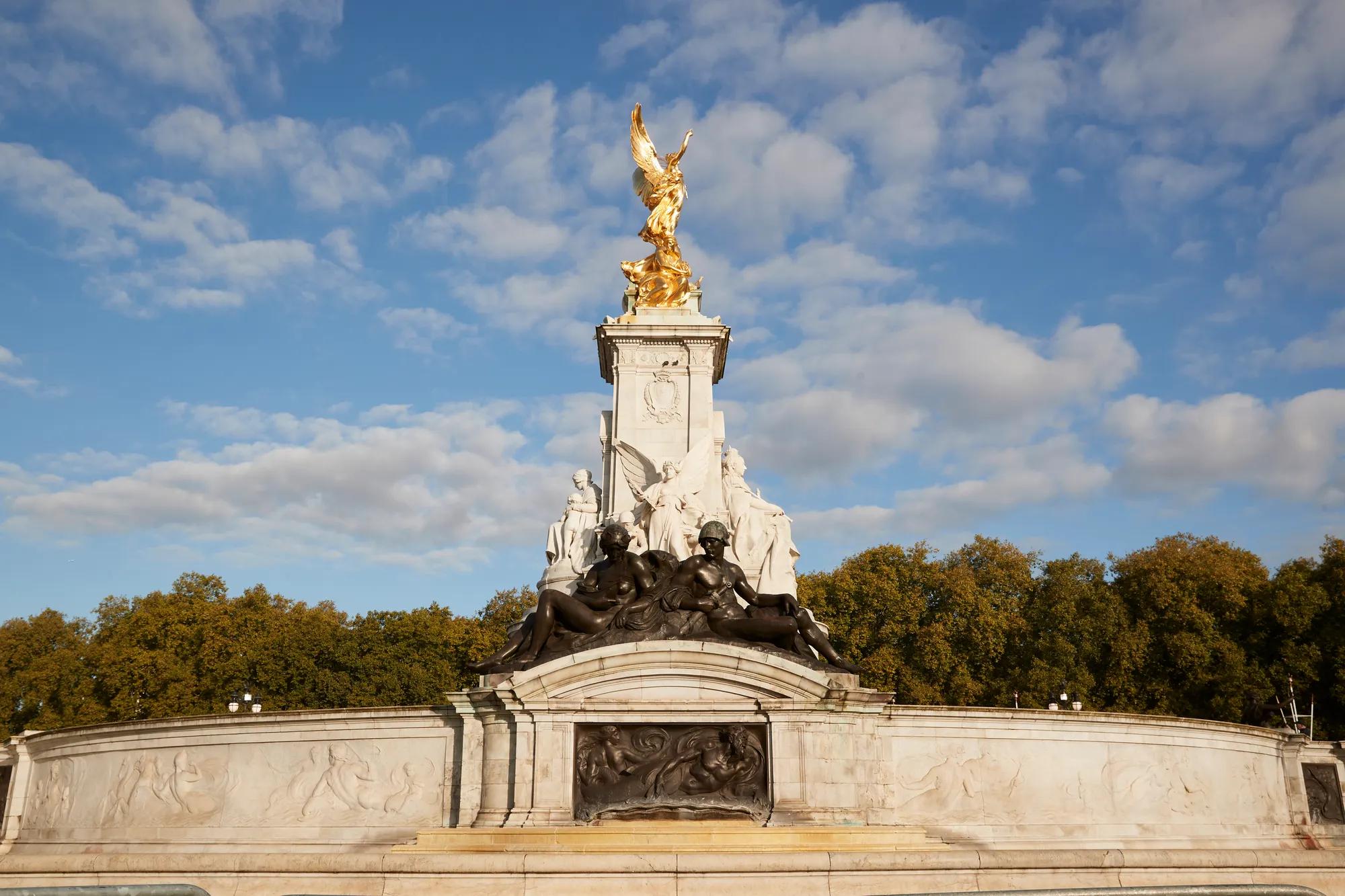 The Victoria Memorial in London, featuring gilded statues atop a white marble monument surrounded by sculpted bronze figures, set against a backdrop of trees and a bright blue sky with scattered clouds.