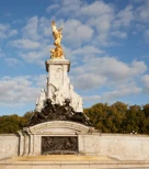 The Victoria Memorial in London, featuring gilded statues atop a white marble monument surrounded by sculpted bronze figures, set against a backdrop of trees and a bright blue sky with scattered clouds.