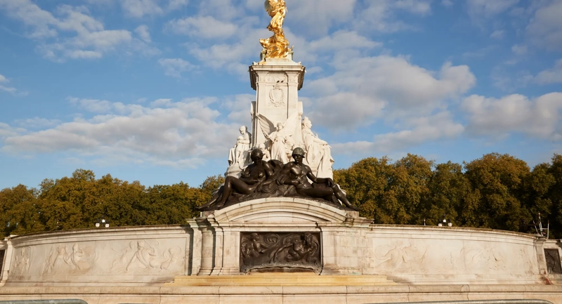 The Victoria Memorial in London, featuring gilded statues atop a white marble monument surrounded by sculpted bronze figures, set against a backdrop of trees and a bright blue sky with scattered clouds.