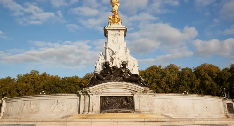 The Victoria Memorial in London, featuring gilded statues atop a white marble monument surrounded by sculpted bronze figures, set against a backdrop of trees and a bright blue sky with scattered clouds.