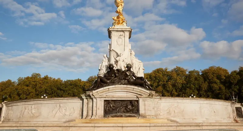 The Victoria Memorial in London, featuring gilded statues atop a white marble monument surrounded by sculpted bronze figures, set against a backdrop of trees and a bright blue sky with scattered clouds.