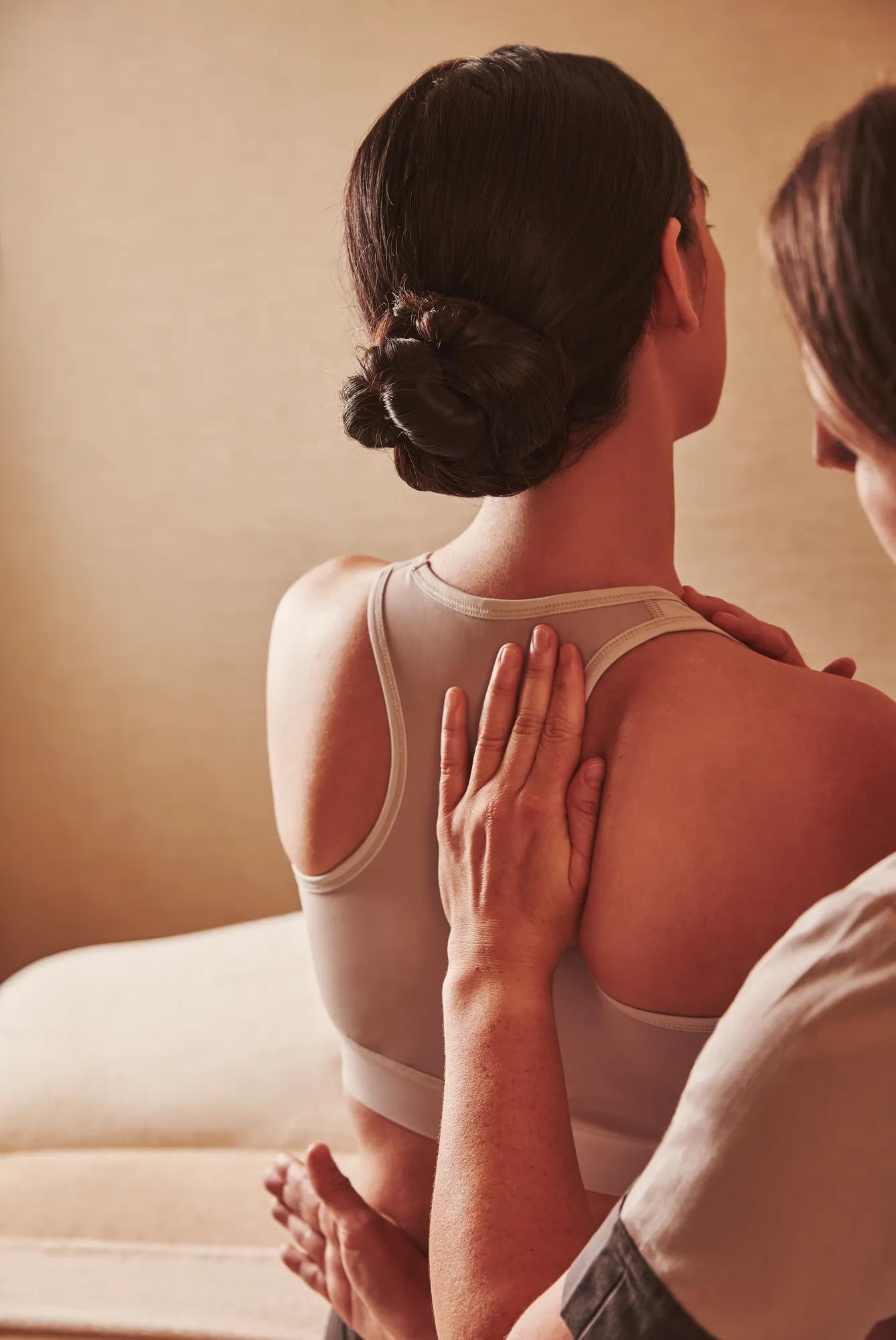 Therapist placing hands on a person's upper back during a seated spa treatment, with the person wearing a sports bra.