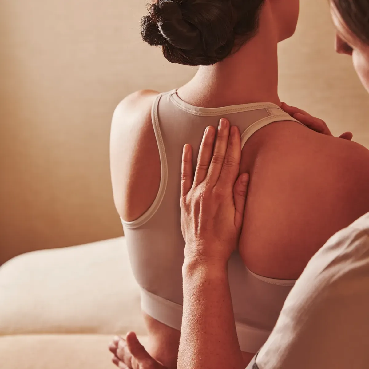Therapist placing hands on a person's upper back during a seated spa treatment, with the person wearing a sports bra.