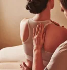 Therapist placing hands on a person's upper back during a seated spa treatment, with the person wearing a sports bra.