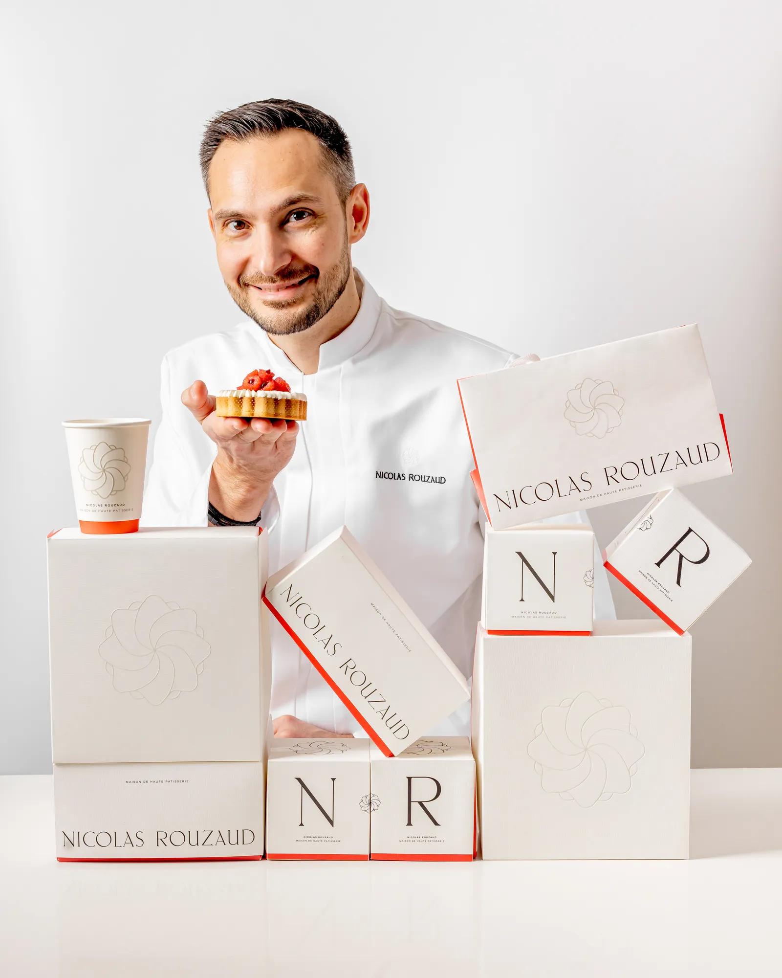Pastry chefNicolas Rouzaud  holding small tart beside stacked Nicolas Rouzaud boxes, smiling at camera against white background.