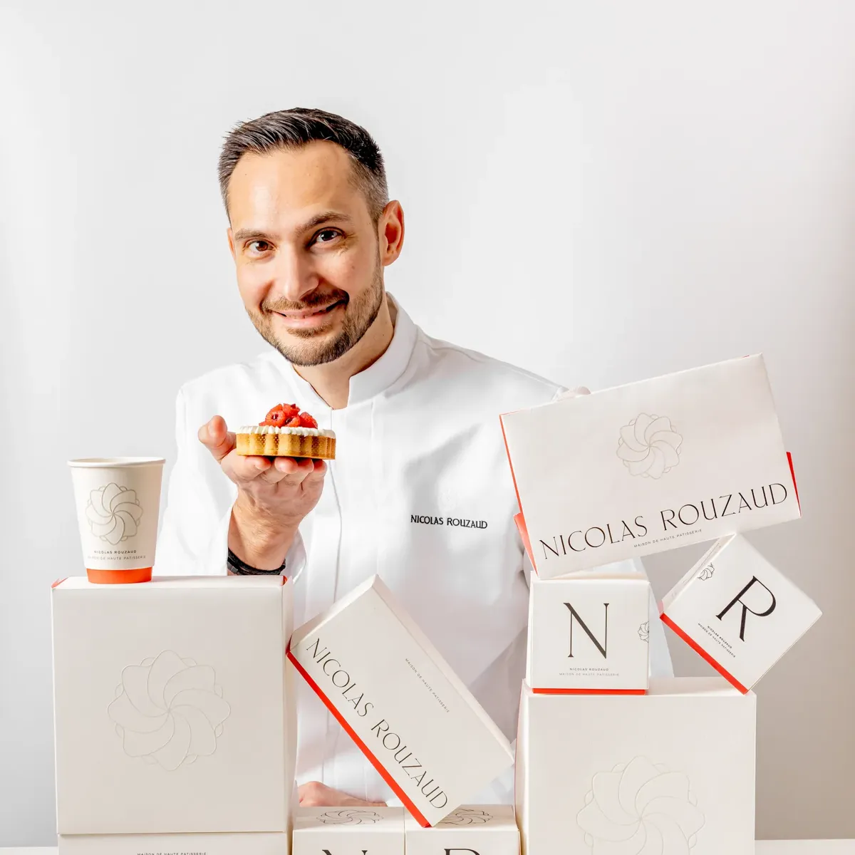 Pastry chefNicolas Rouzaud  holding small tart beside stacked Nicolas Rouzaud boxes, smiling at camera against white background.