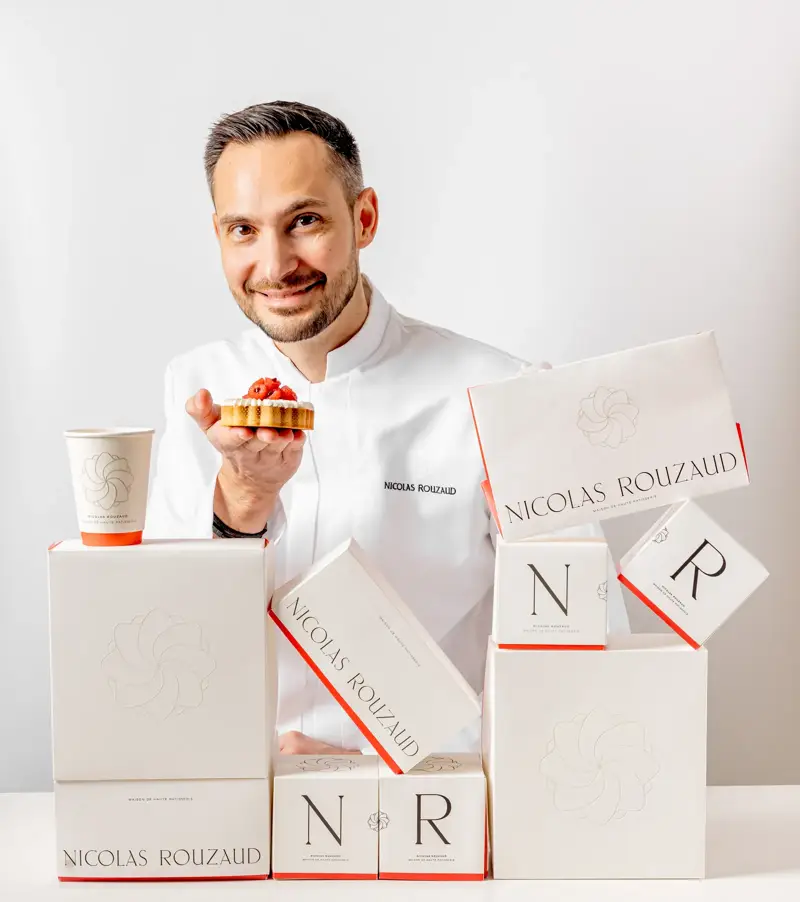 Pastry chefNicolas Rouzaud  holding small tart beside stacked Nicolas Rouzaud boxes, smiling at camera against white background.