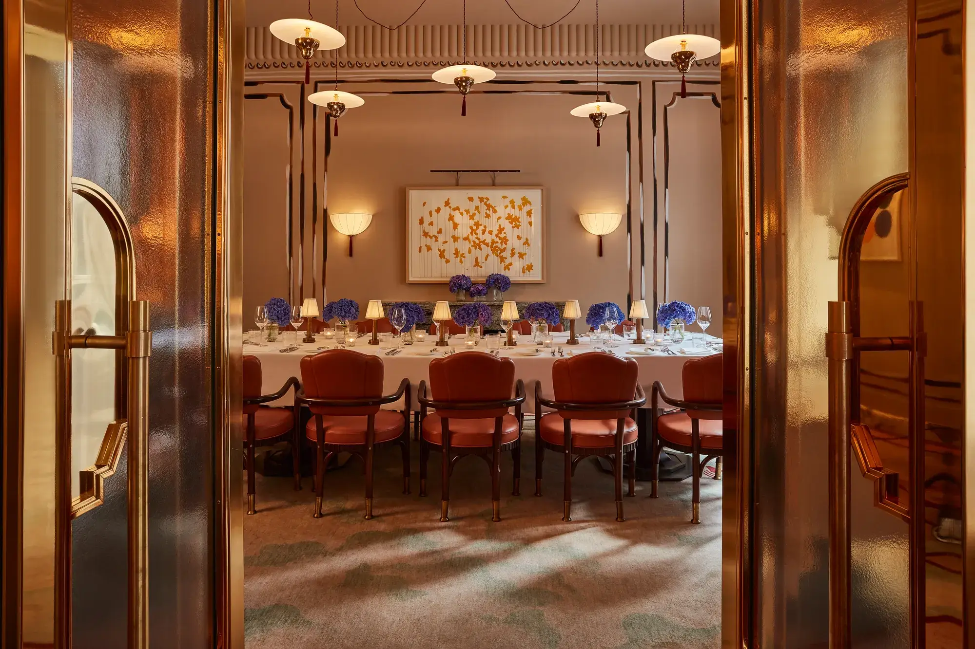Elegant private dining room viewed through golden double doors, featuring a long table set with white linens, blue hydrangea centerpieces, and soft lamp lighting. Leather chairs and contemporary wall art create a refined, intimate ambiance.
