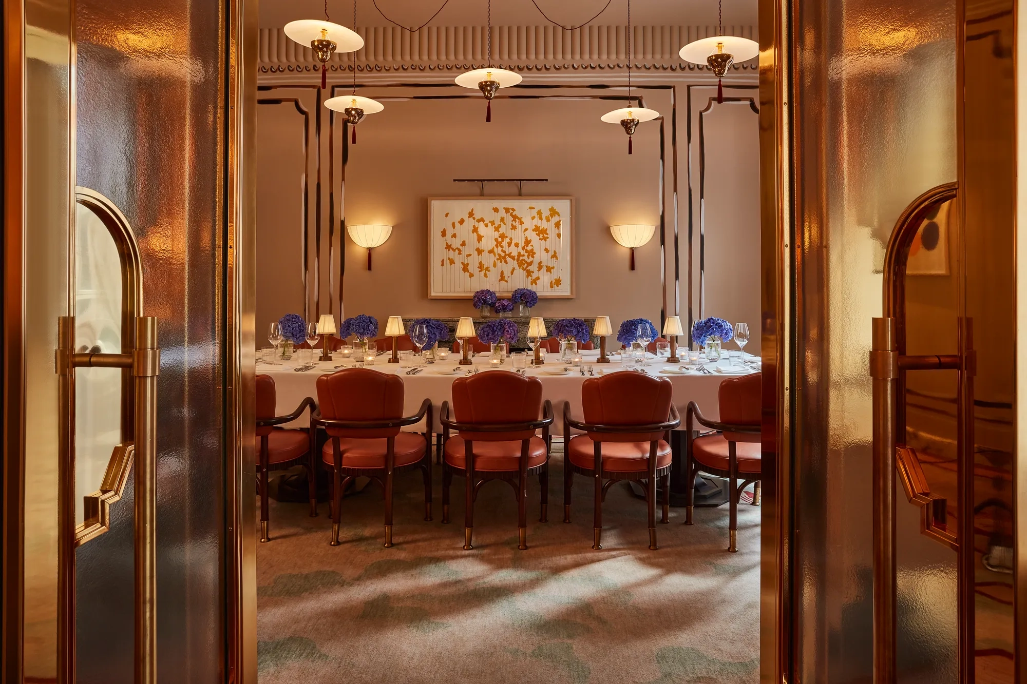 Elegant private dining room viewed through golden double doors, featuring a long table set with white linens, blue hydrangea centerpieces, and soft lamp lighting. Leather chairs and contemporary wall art create a refined, intimate ambiance.