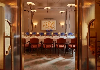 Elegant private dining room viewed through golden double doors, featuring a long table set with white linens, blue hydrangea centerpieces, and soft lamp lighting. Leather chairs and contemporary wall art create a refined, intimate ambiance.