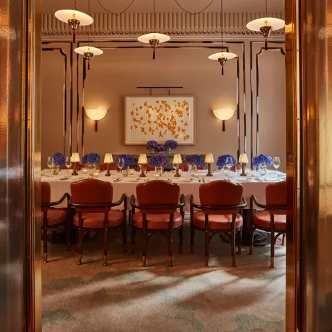 Elegant private dining room viewed through golden double doors, featuring a long table set with white linens, blue hydrangea centerpieces, and soft lamp lighting. Leather chairs and contemporary wall art create a refined, intimate ambiance.