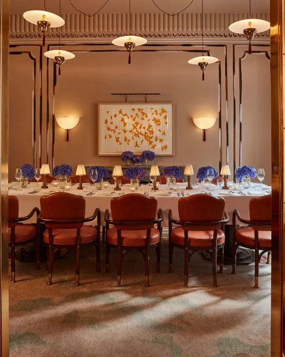 Elegant private dining room viewed through golden double doors, featuring a long table set with white linens, blue hydrangea centerpieces, and soft lamp lighting. Leather chairs and contemporary wall art create a refined, intimate ambiance.