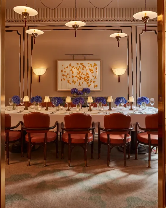Elegant private dining room viewed through golden double doors, featuring a long table set with white linens, blue hydrangea centerpieces, and soft lamp lighting. Leather chairs and contemporary wall art create a refined, intimate ambiance.