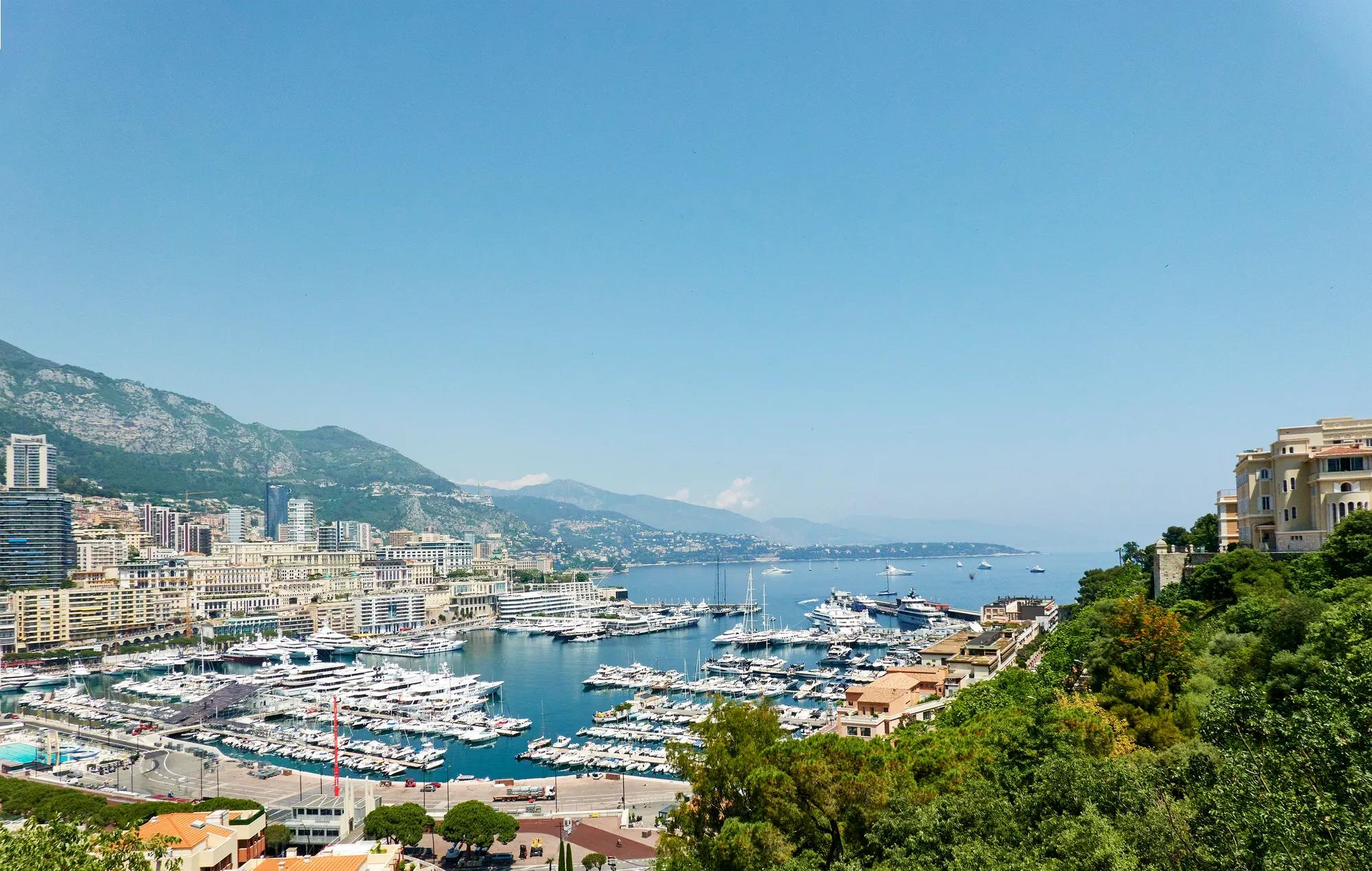 View overlooking Monaco’s marina with yachts, coastal buildings, and mountains under a clear blue sky.