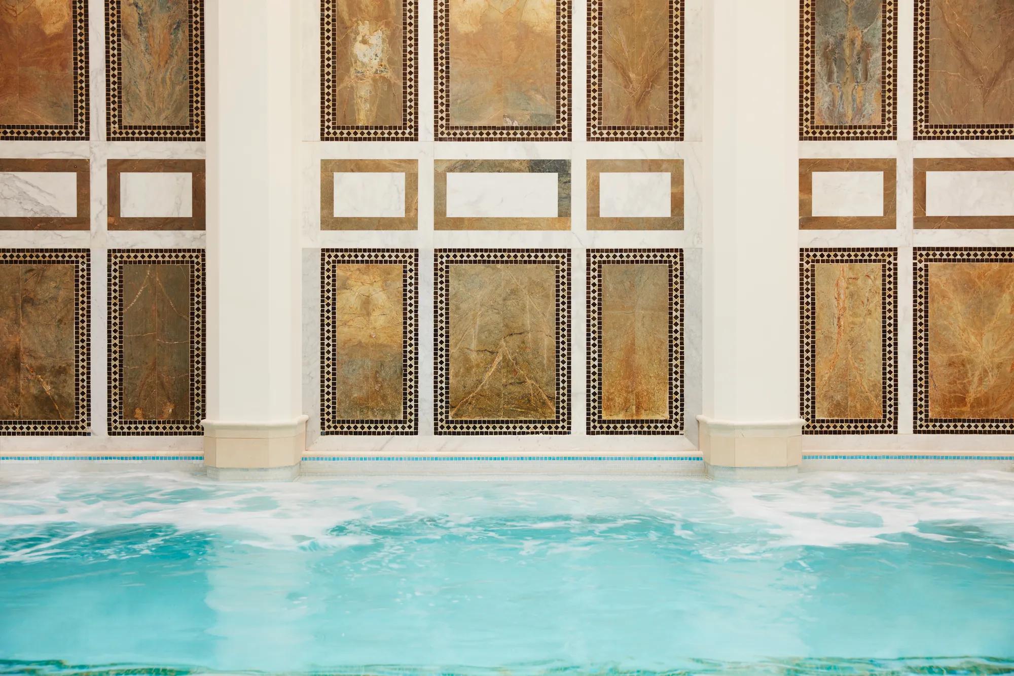 Close-up view of an indoor spa pool with gently bubbling turquoise water in the foreground. The wall behind the pool features symmetrical rectangular stone panels in warm gold and earthy tones, framed by intricate mosaic tile borders and white marble accents between columns, creating an elegant, geometric design.