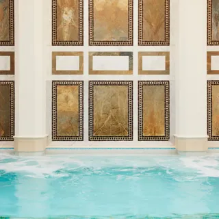 Close-up view of an indoor spa pool with gently bubbling turquoise water in the foreground. The wall behind the pool features symmetrical rectangular stone panels in warm gold and earthy tones, framed by intricate mosaic tile borders and white marble accents between columns, creating an elegant, geometric design.